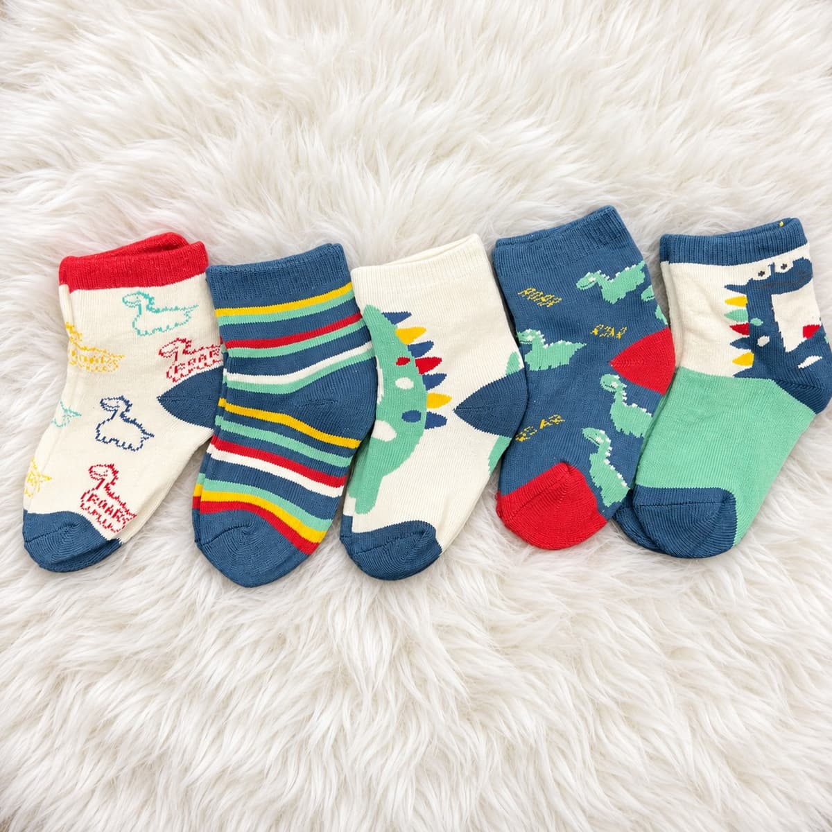 Five boys' dinosaur socks on a white rug: cream with outline dinosaurs and 'ROAR', navy stripes, cream with a green spiky dinosaur, navy with green dinosaurs and red heels, and cream and green with a navy dinosaur