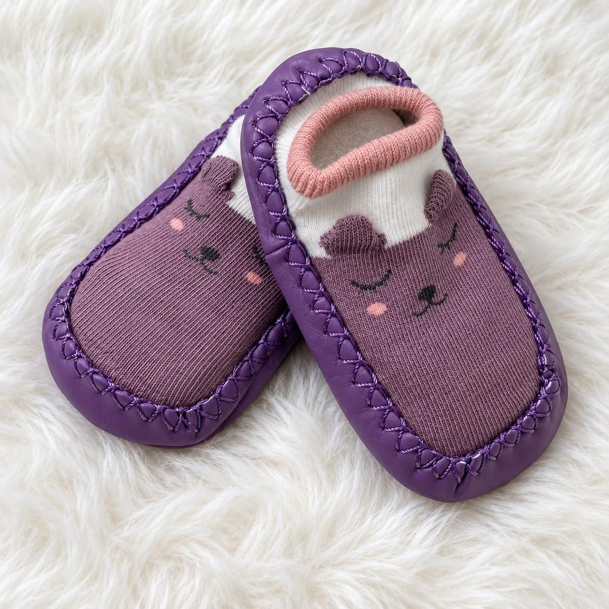Purple bear sock shoes with a sleepy face, small ears and a coral-rimmed cuff, on white fur