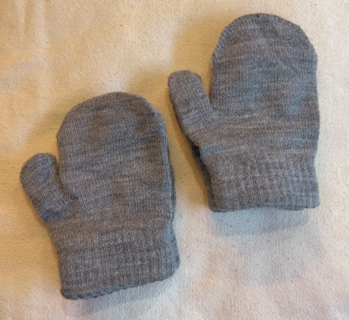 Mid grey knitted baby mittens with ribbed cuffs, laid flat on cream cloth