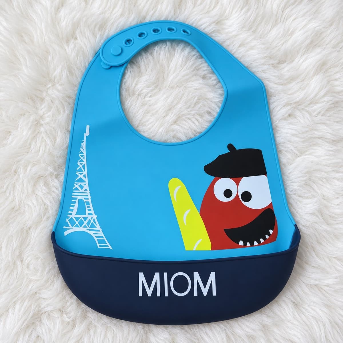 Blue MIOM silicone bib with a navy crumb pocket, printed with the Eiffel Tower and a red character in a beret holding a baguette, on white fur